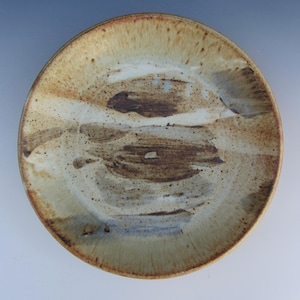 May include: A round ceramic plate featuring a speckled beige and cream base with prominent, wide, horizontal brushstrokes in dark brown and muted gray across the center. The rim is defined by a darker, toasted brown edge with fine speckling.