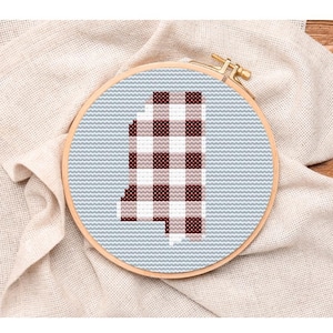 May include: A cross-stitch embroidery hoop featuring the state of Mississippi. The state outline is filled with a red and white gingham pattern, set against a light blue background. The hoop is made of wood and rests on a beige fabric.