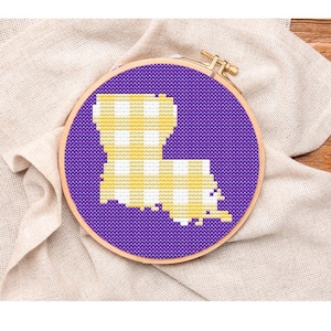 May include: A cross-stitch embroidery hoop featuring the state of Louisiana in a purple background. The state is filled with a yellow and white gingham pattern. The hoop is made of wood and is set against a neutral fabric.