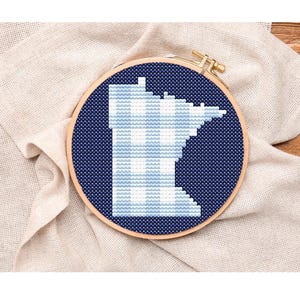 May include: A cross-stitch embroidery hoop featuring the outline of the state of Minnesota in light blue and white against a dark blue background. The design is set in a wooden embroidery hoop, resting on a beige linen cloth.