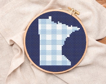 May include: A cross-stitch embroidery hoop featuring the outline of the state of Minnesota in light blue and white against a dark blue background. The design is set in a wooden embroidery hoop, resting on a beige linen cloth.