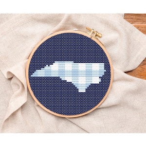 May include: A cross-stitch embroidery hoop featuring the outline of North Carolina in light blue and white against a dark blue background. The embroidery is set in a wooden hoop and rests on a beige fabric.