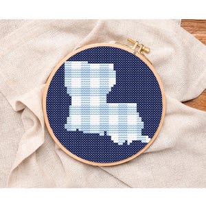 May include: A cross-stitch embroidery hoop featuring the outline of the state of Louisiana in a blue and white gingham pattern against a dark blue background. The embroidery is set in a wooden hoop, resting on a beige linen cloth.