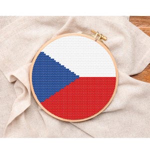 Pode incluir: Um bastidor de bordado em ponto cruz com a bandeira da República Tcheca. O design inclui as cores azul, branco e vermelho. O bastidor está sobre um fundo de tecido bege e uma superfície de madeira.