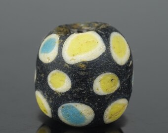 May include: A black bead with a series of yellow and blue oval-shaped designs. The bead has a cylindrical shape with a hole running through the center. The bead's surface has a slightly rough texture.