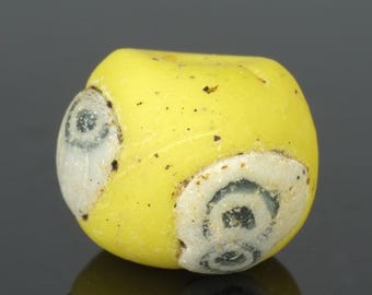 May include: A yellow glass bead with white and black swirl designs. The bead is round and has a smooth surface.