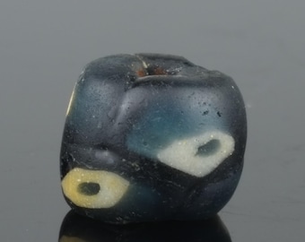 May include: A dark blue, rounded bead with white and yellow eye-like designs. The bead has a slightly irregular shape and a glossy finish, suggesting it may be made of glass or a similar material. The eye-like designs are oval-shaped.