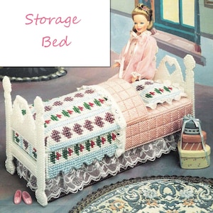 May include: A white plastic doll bed with a pink and white quilt, a white pillow with green and red accents, and a white lace trim. The bed has a heart-shaped headboard and footboard. The bed is sitting on a blue and white rug. The text "Storage Bed" is written in pink letters on a white background.