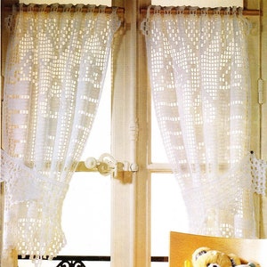 May include: Two white crocheted curtains hang in a window with a black wrought iron railing. A wicker basket with a teddy bear, a baby bottle, and a blue and white stuffed animal sits on a windowsill.