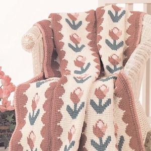 Puede incluir: Una manta de ganchillo con un patrón floral en tonos rosa, marrón y verde. La manta está colocada sobre una silla de mimbre blanca.