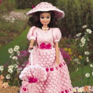 Peut inclure: Une robe de poupée en tricot rose avec une bordure en dentelle blanche et un ruban de satin rose. La poupée porte un chapeau blanc avec une bordure en tricot rose. La poupée tient un sac à main en tricot rose.