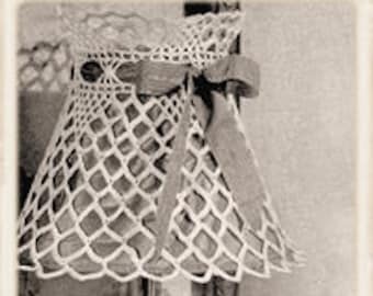 Puede incluir: Una pantalla de lámpara de crochet blanco con un gran lazo atado. La pantalla está sobre una base de lámpara plateada con una base de vidrio transparente.