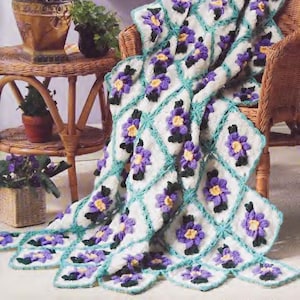 Peut inclure: Une couverture en crochet avec un motif floral dans les tons de violet, jaune et vert. La couverture est posée sur une chaise en osier et il y a des plantes en pot en arrière-plan.