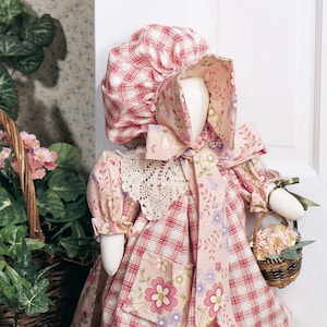 Op de afbeelding: Een stoffen pop in een roze geruite jurk met bloemaccenten, een bijpassende muts en een kanten kraag. De pop draagt een klein mandje met roze bloemen, wat een vintage, landelijke uitstraling creëert.