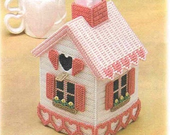 May include: A pink and white tissue box cover shaped like a house with a heart-shaped window and red shutters. The roof has a pink and white checkered pattern and the base is decorated with pink hearts.
