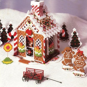 May include: A gingerbread house with candy cane trim, a red roof, and a white picket fence. Two gingerbread people stand in front of the house, and a small red wagon sits in the snow.