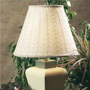 May include: A white lampshade with a delicate lace pattern sits atop a cream-colored ceramic lamp base. The lamp is on a table with green plants in pots.