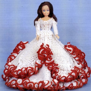 Op de afbeelding: Een witte gehaakte poppenjurk met rode ruches. De jurk heeft een kantboord en een volle rok.