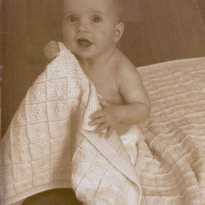 May include: A baby is sitting on a white knitted blanket. The blanket has a textured pattern and is made of soft yarn.