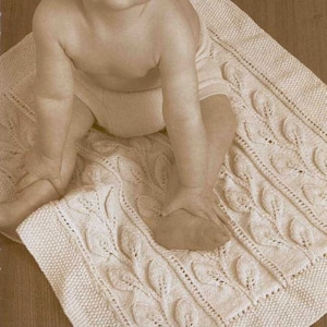 May include: A baby sitting on a white knitted blanket with a leaf pattern. The blanket is made with a cable knit stitch.