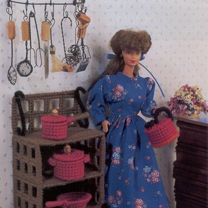 以下が含まれることがあります： 青い花柄のドレスを着た人形が赤い編み物のバスケットを持っています。人形は赤い編み物の鍋やフライパンが置かれた茶色の木製の棚の前に立っています。