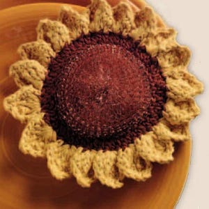 May include: A crocheted sunflower with a brown center and yellow petals. The sunflower is sitting on a brown plate.