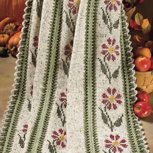 Könnte beinhalten: Eine gehäkelte Wolldecke mit weißem Hintergrund und grünen und roten Blumendesigns. Die Decke hat einen gehäkelten Rand mit einem gewellten Saum.