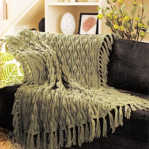 May include: A sage green knitted throw blanket featuring a repeating textured leaf pattern throughout the fabric. The rectangular blanket is finished with long, matching green fringe tassels along the edges, creating a decorative border.