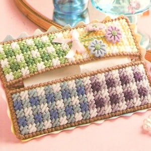 Puede incluir: Una funda para caja de pañuelos hecha a mano con un patrón de cuadros verde, blanco y morado. La solapa superior presenta un diseño floral amarillo y blanco con una cinta rosa.