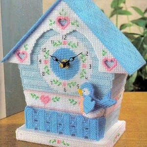 Può includere: Un orologio a forma di casetta per uccelli blu e bianco con un uccello blu e cuori rosa. Il quadrante dell'orologio ha uno sfondo blu con numeri e lancette bianche.