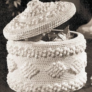May include: A white crocheted storage box with a lid. The box has a textured design with raised bumps and a circular shape. The lid is slightly domed and has a similar textured design.