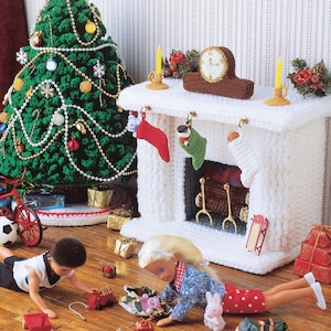 May include: A white crocheted fireplace with a clock and stockings hanging on it. Two dolls are playing with toys on a wooden floor in front of a green crocheted Christmas tree.