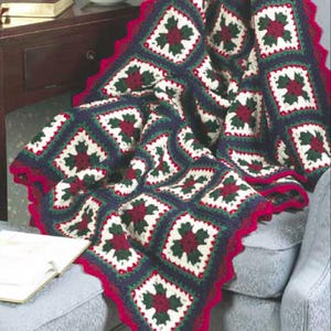 May include: A handmade crochet blanket with a festive design. The blanket features square motifs in navy blue, white, green, and burgundy, with a scalloped red border. The blanket is draped over a chair.