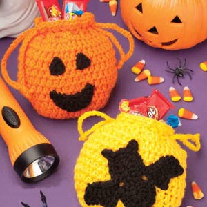May include: Two crocheted Halloween treat bags, one orange with a jack-o'-lantern face and the other yellow with a black bat. Candy corn and a plastic pumpkin and ghost are also visible.