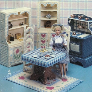 Op de afbeelding: Een poppenhuis-keukenset met een witte kast, een hoekkast en een blauw fornuis. De set bevat een tafel met een geruit tafelkleed, twee stoelen en een pop.