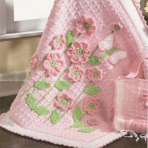 May include: A pink crocheted baby blanket with a white border and a floral design. The blanket features green leaves and pink flowers.