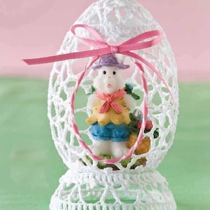 May include: A white crocheted Easter egg with a pink ribbon bow. Inside, a small figurine of a bunny wearing a yellow vest, blue shorts, and a purple hat is visible. The egg is on a white crocheted base.