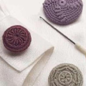 Puede incluir: Tres botones de ganchillo en diferentes colores: morado, marrón y gris. Cada botón tiene un diseño único en el centro.
