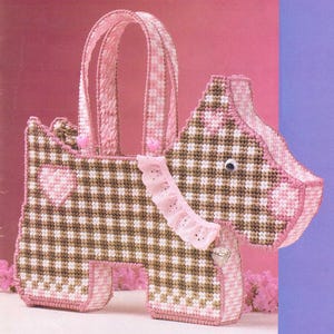 Puede incluir: Una bolsa de mano con forma de perro de cuadros marrones y rosas con un corazón rosa en la parte delantera y un ribete de volantes rosa. La bolsa tiene un asa de cuadros rosa y blancos.