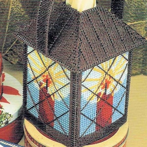 Puede incluir: Una funda para caja de pañuelos de color marrón y azul con forma de linterna con una vela en su interior. La linterna tiene una ventana de vidrio transparente y un marco de metal marrón. La vela es roja y tiene una llama amarilla.