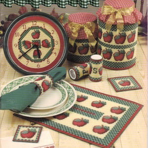 May include: A collection of needlepoint kitchen accessories featuring red apple motifs and green checkered borders. The set includes a round wall clock, two lidded canisters with gold ribbons, a napkin ring, salt and pepper shakers, a placemat, and two square coasters.