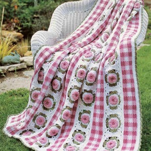 May include: A handmade crochet blanket draped over a white wicker chair. The blanket features a pattern of pink gingham stripes and white squares with pink flower motifs. The edges are trimmed with a green border.