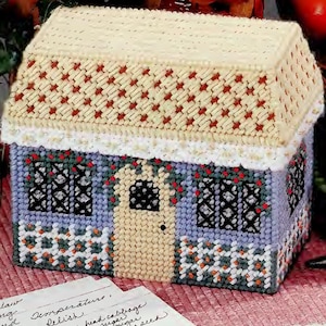 May include: A plastic canvas craft house featuring a cream-coloured roof with red accents, light blue walls, a central cream door, black lattice windows with red floral details, and a white picket fence with orange flowers along the base.