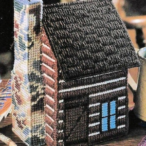 May include: A plastic canvas craft project shaped like a small cabin with a dark brown roof and walls, white horizontal accents, and a blue window. Attached to the side is a tall, rectangular chimney featuring a multicolored geometric pattern.
