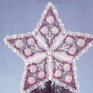 May include: A white star-shaped Christmas tree topper with a snowflake design in the center. The snowflake is made of white and gray yarn and is surrounded by pink and purple yarn flowers. The star is trimmed with white lace.