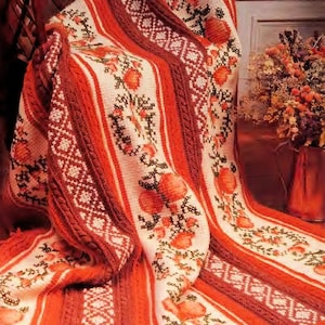 May include: A handmade, knitted blanket with a floral pattern. The blanket features a cream-colored base with vertical stripes in shades of red and brown. The floral design includes red flowers and green leaves, with geometric patterns along the edges.