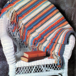 May include: A striped crocheted throw blanket with fringe in shades of blue, red, white and beige. The blanket is draped over a white wicker chair with two books on the seat.