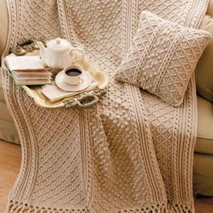May include: A crocheted throw blanket with a diamond pattern and a fringe border. The blanket is beige and has a textured surface. A matching crocheted pillow is also shown.