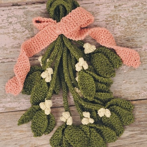 May include: A handmade knitted mistletoe decoration with green leaves and white flowers, tied with a pink ribbon.