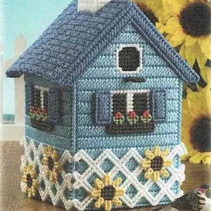 Puede incluir: Una funda para caja de pañuelos de tejido azul y blanco con forma de casa con una valla blanca y girasoles alrededor de la base.
